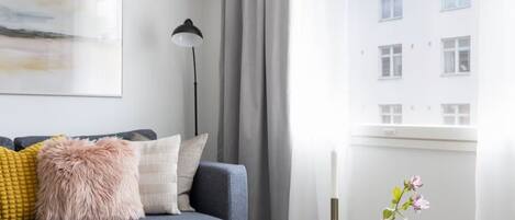 A modern living room with a grey sofa, a round ottoman, a small table with a candle and flowers, and a large window with curtains.