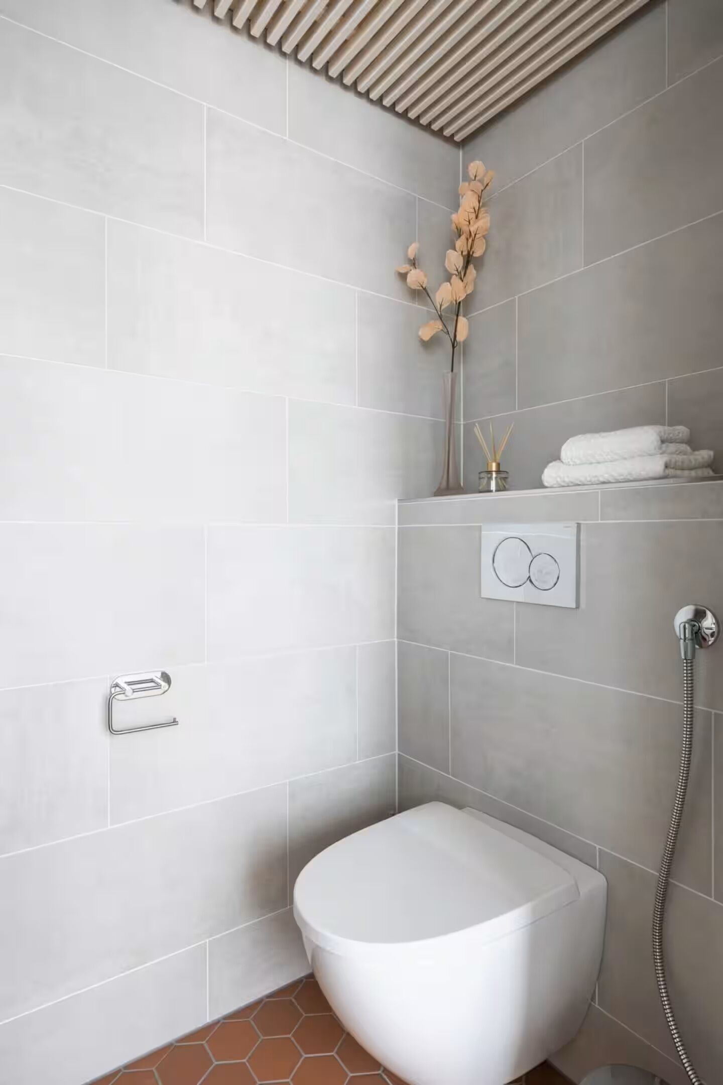 A modern bathroom with grey tiles, a white toilet, a shower with a handheld showerhead, and a decorative plant.