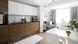 A modern kitchen with wooden cabinets, a black countertop, and a built-in oven. There is a dining area with a white table and chairs, and a gray sofa with decorative pillows. - New Stylish 1BR Apt. in Trendy Kallio (Helsinki)