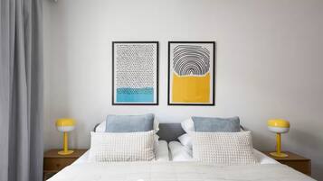Un lit bien fait, avec du linge de lit blanc et deux oreillers, est encadré de deux tables de chevet sur lesquelles reposent deux lampes jaunes. Deux œuvres d’art abstraites, encadrées, sont accrochées au mur au-dessus du lit.