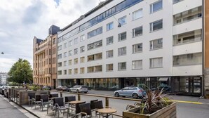 A modern hotel with a row of rooms on the ground floor, outdoor seating area with tables and chairs, and a parking lot with cars. - New Stylish 1BR Apt. in Trendy Kallio (Helsinki)