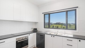 A modern kitchen with white cabinets, a black countertop, a dishwasher, and an oven. A large window provides a view of a building and greenery outside.