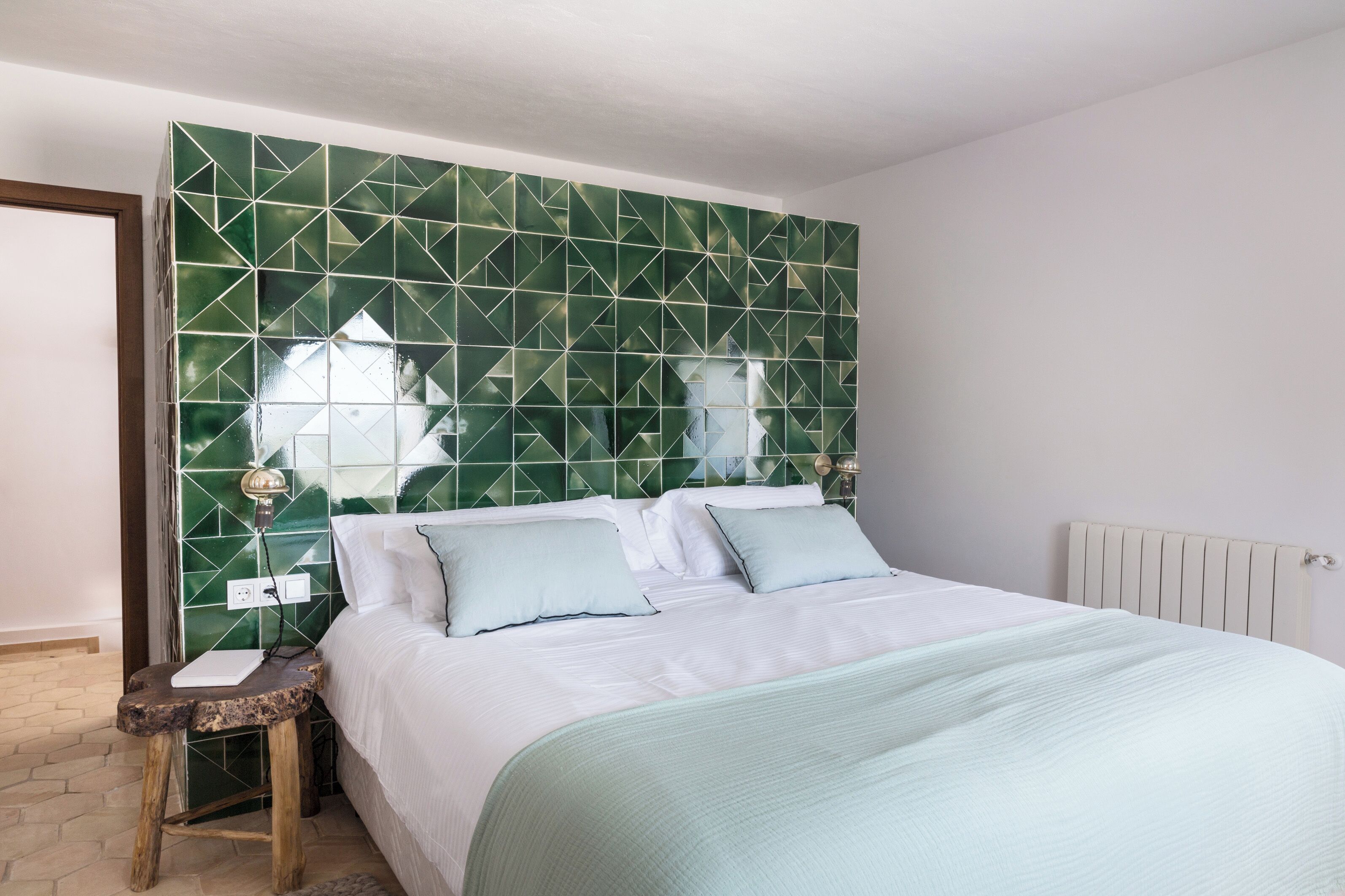 Une chambre avec un mur orné d’un motif géométrique vert, un lit blanc avec une literie bleu clair et une table de chevet en bois avec une lampe.