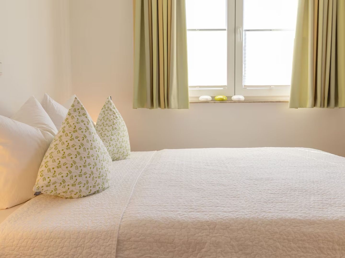 Ein ordentlich bezogenes Bett mit weißen und grün gemusterten Kissen, einer weißen Decke und einem Fenster mit grünen Vorhängen.