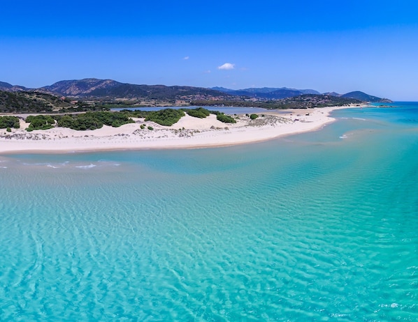 Et kystlandskab med en sandstrand, krystalklart turkis vand og fjerne bjerge.