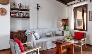 Una sala de estar con un sofá, dos sillones, una mesa de centro de madera, una estantería, una lámpara y una ventana con cortinas.