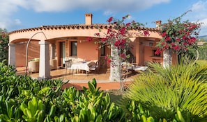 A building with a patio, outdoor seating, and a garden with plants and flowers. - Dream Villa with Panoramic Terrace near Costa Rei Beach (Costa Rei)