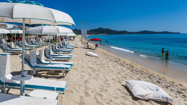 A beach with rows of sun loungers and umbrellas, a clear blue sky, and a calm sea.