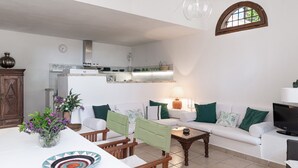 A living room with a sofa, chairs, a coffee table, a TV, and a kitchen area with cabinets and a window. - Villa Fiori 4 Is Molas – Exclusive Villa with Private Pool and Sea View in Sardinia (S.Margh.di Pula)