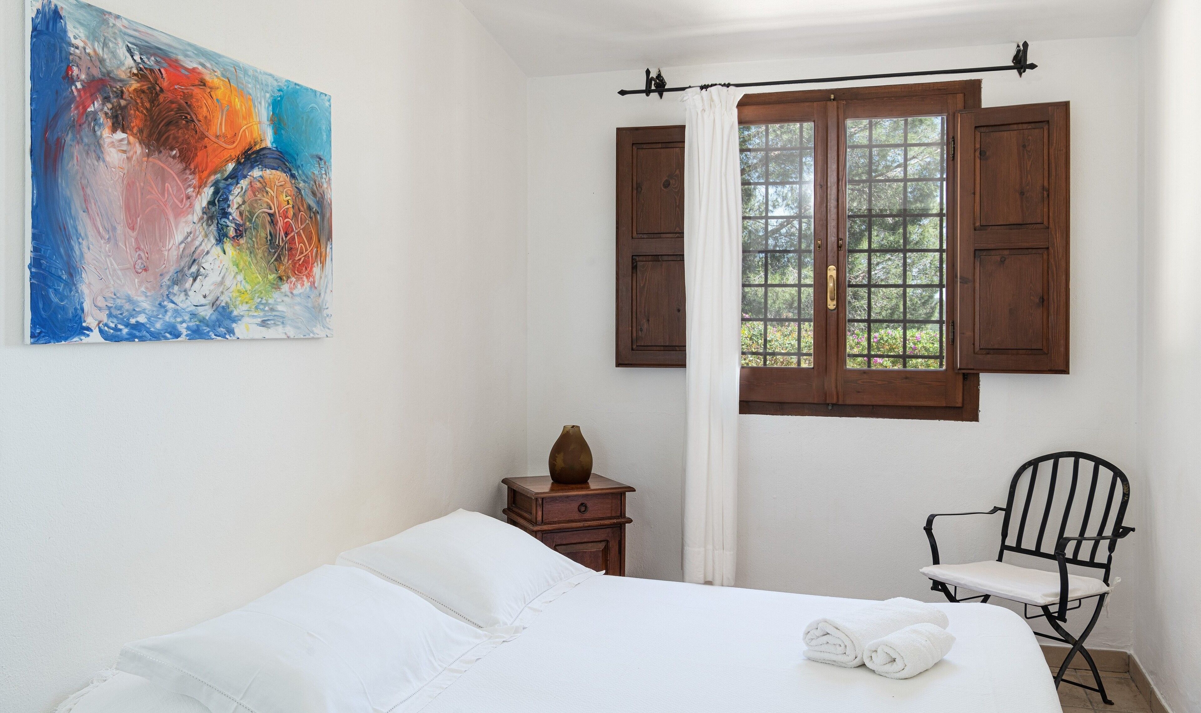 A bedroom with a bed, a chair, a nightstand, a window with shutters, and a painting on the wall.