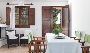 A dining area with a table set for two, two wicker chairs, a wooden door, and a window with shutters.