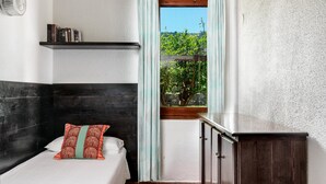 A bedroom with a bed, a nightstand, a shelf with books, a window with a view of greenery, and a wooden cabinet. - Villa Roberta at Costa Rei – Sardinian Beach Villa with Garden (Costa Rei)