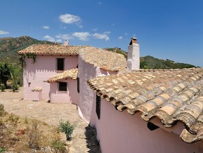 A pink building with a tiled roof, a small garden, and a mountainous backdrop. - Finca Fiori 2 – Peaceful Country House with Private Pool in Sardinia (S.Margh.di Pula)