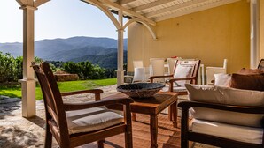 A covered outdoor seating area with a view of mountains, wooden furniture, and a stone table. - Villa Aquila Giardino – Nature Retreat with Panoramic Views and Private Pool (S. Margh. di Pula)