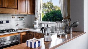 A kitchen with wooden cabinets, a stove, a coffee maker, and a window with a view of greenery. - Villa Aquila Giardino – Nature Retreat with Panoramic Views and Private Pool (S. Margh. di Pula)