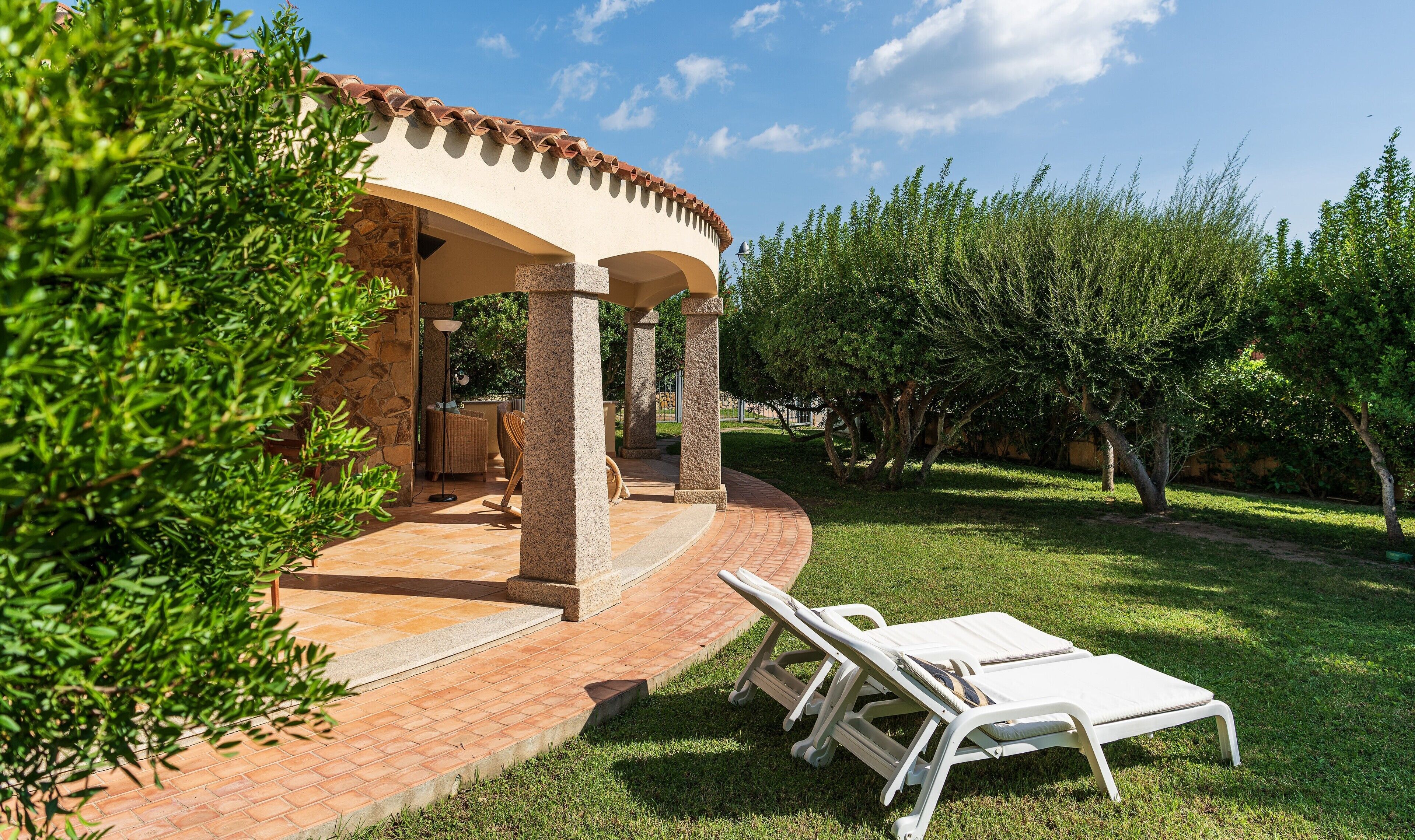 A covered outdoor seating area with stone pillars, a brick pathway, and a white lounge chair.