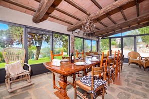 A dining area with a wooden table set for four, surrounded by wicker chairs with cushions, large windows offering a view of greenery, and a chandelier hanging from the ceiling. - Casale Mulino (Scansano)