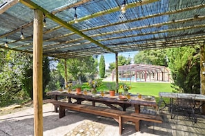 An outdoor patio with a covered seating area, a table set for dining, and a pool in the background. - Casale Mulino (Scansano)