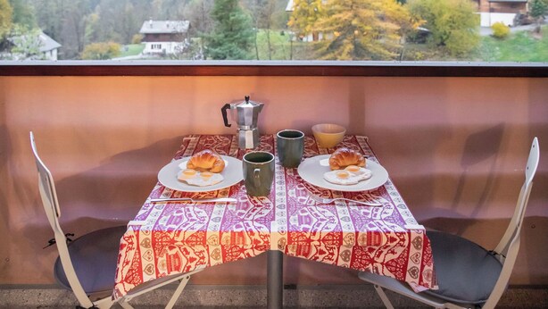 Ein Tisch mit gemustertem Tischläufer, zwei Teller mit Croissants und Eiern, eine Kaffeekanne und eine Tasse, vor einer malerischen Bergkulisse.