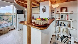 Una cocina con techo de madera, una cesta de frutas sobre un mostrador de madera y una estantería blanca con diversos objetos.