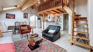 Un salón amplio con techo alto, vigas de madera, un sofá de cuero negro, un sillón rojo, una mesa de centro de vidrio y una alfombra estampada.