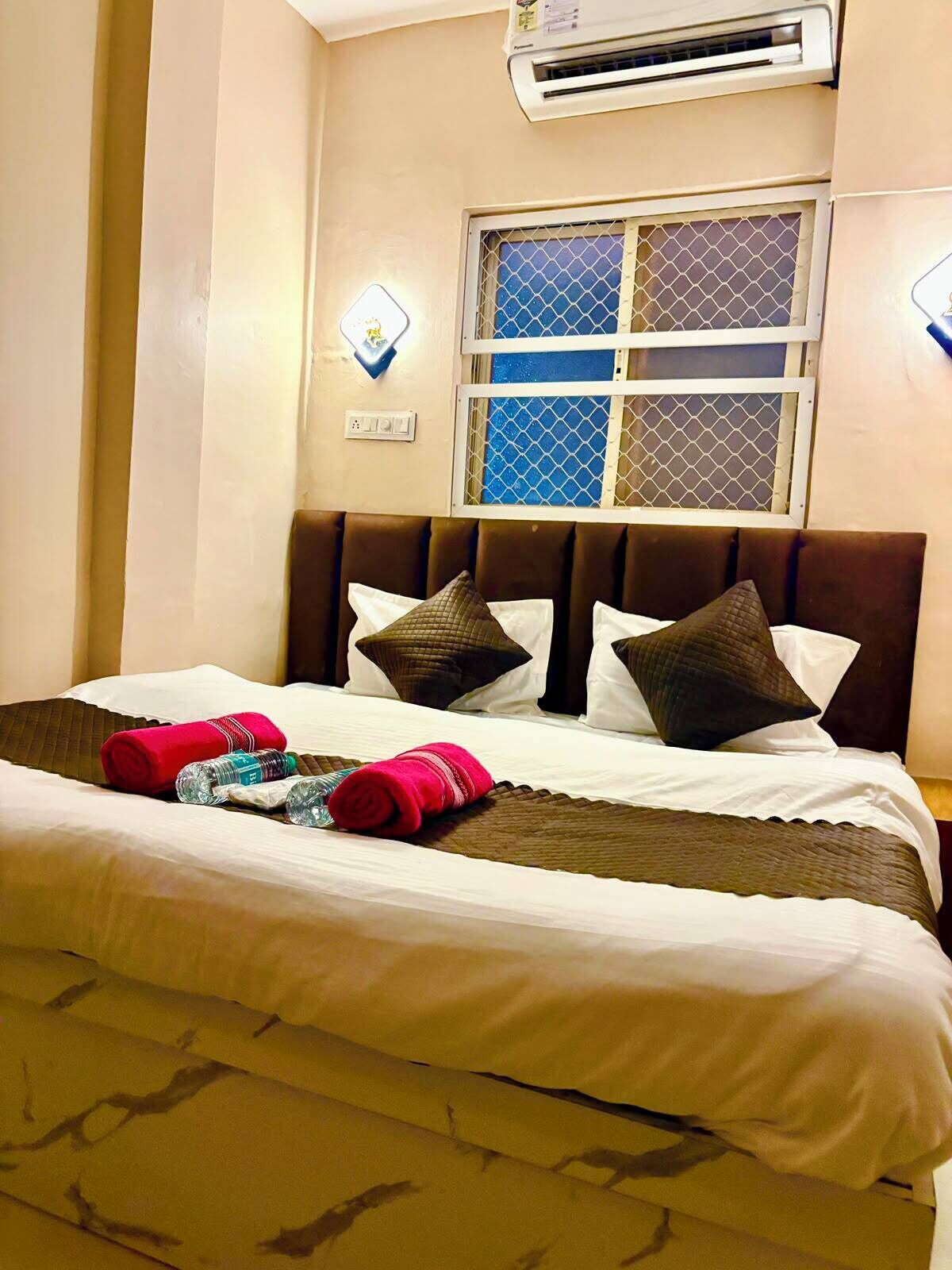 A hotel room with a bed, pillows, a window with a mesh screen, an air conditioner, and wall-mounted lights.