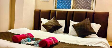 A hotel room with a bed, pillows, a window with a mesh screen, an air conditioner, and wall-mounted lights.