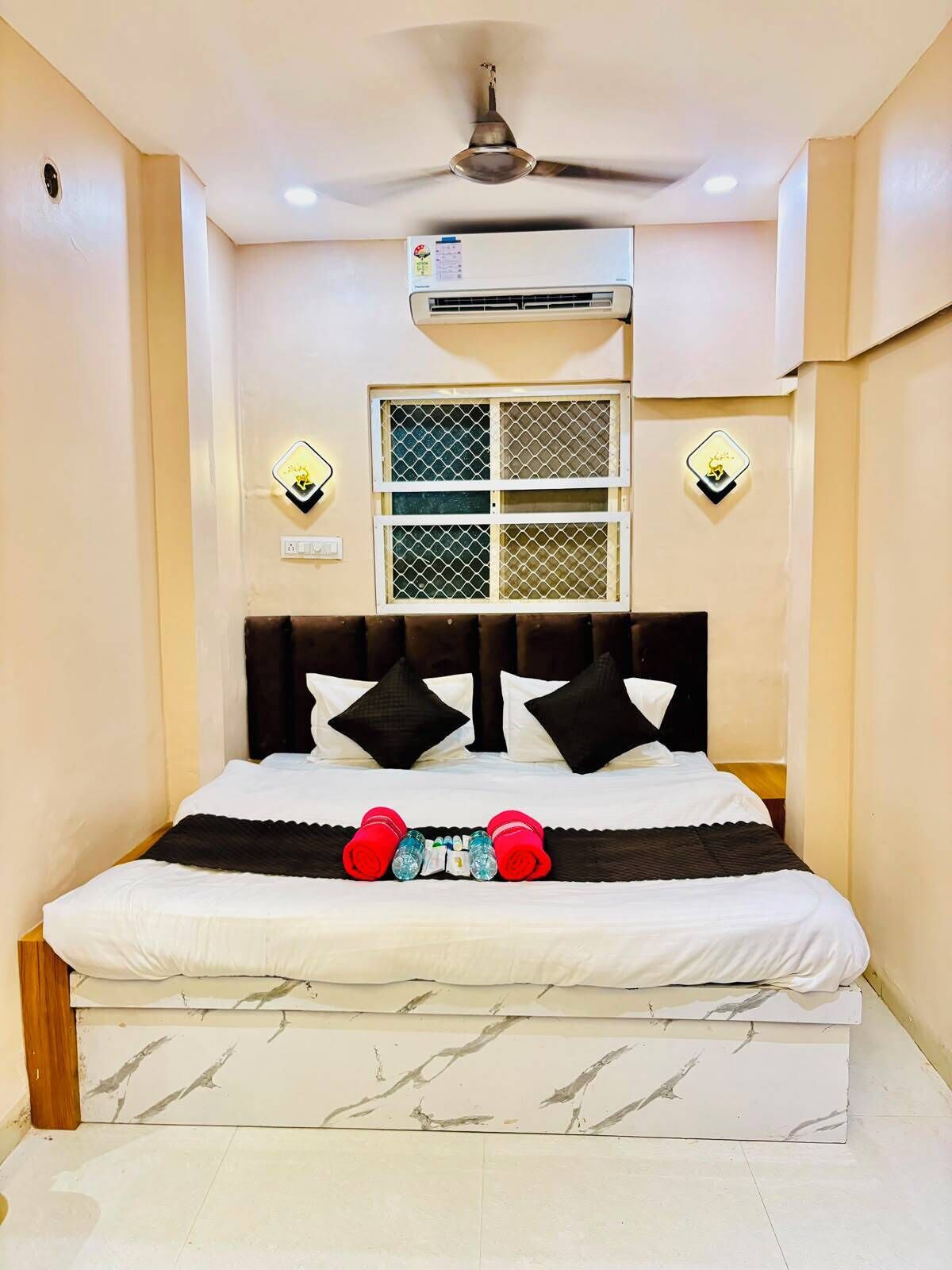 A bedroom with a bed, a ceiling fan, an air conditioning unit, a window with a mesh screen, and wall-mounted light fixtures.