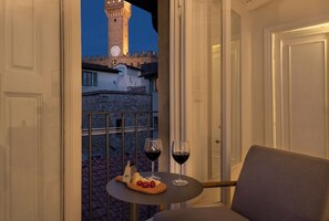 A balcony with a table set for two, featuring a cheese board, tomatoes, and two glasses of red wine. A stone wall and a clock tower are visible in the background.