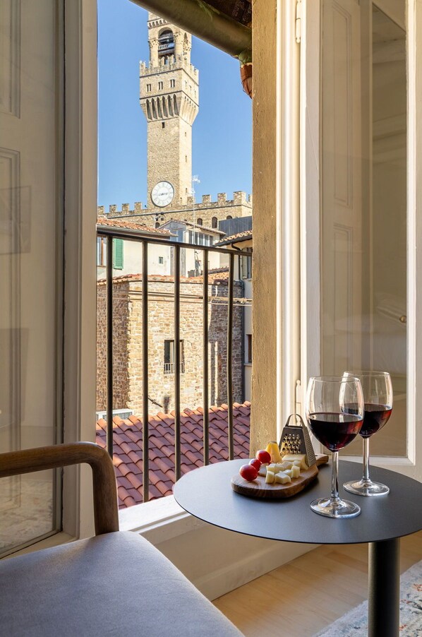 A balcony with a table, two wine glasses, and a cheese board. A historic tower is visible in the background.