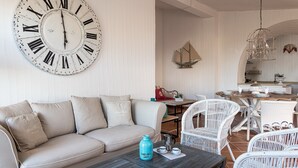 Sala de estar com um relógio de parede grande, um sofá, uma mesa de centro e um espaço de jantar com cadeiras de vime.