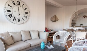 A living room with a large wall clock, a sofa, a coffee table, and a dining area with wicker chairs. - Li Conchi 33 – Family-Friendly Villa with Pool near Cala Sinzias (Castiadas)