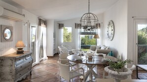 Uma sala de jantar com mesa redonda, cadeiras de vime branco, lustre e vista para o verde lá fora.