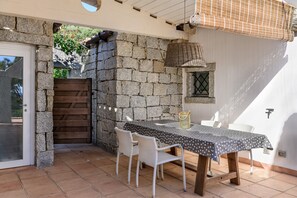 Une terrasse avec un mur en pierre, une porte en bois, une table avec des chaises et un panier suspendu.