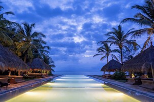 Uma piscina infinita com vista para o oceano, cercada por palmeiras e guarda-sóis de palha.