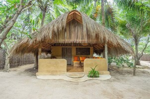 Uma cabana com telhado de palha e chão de areia, rodeada por palmeiras e vegetação.
