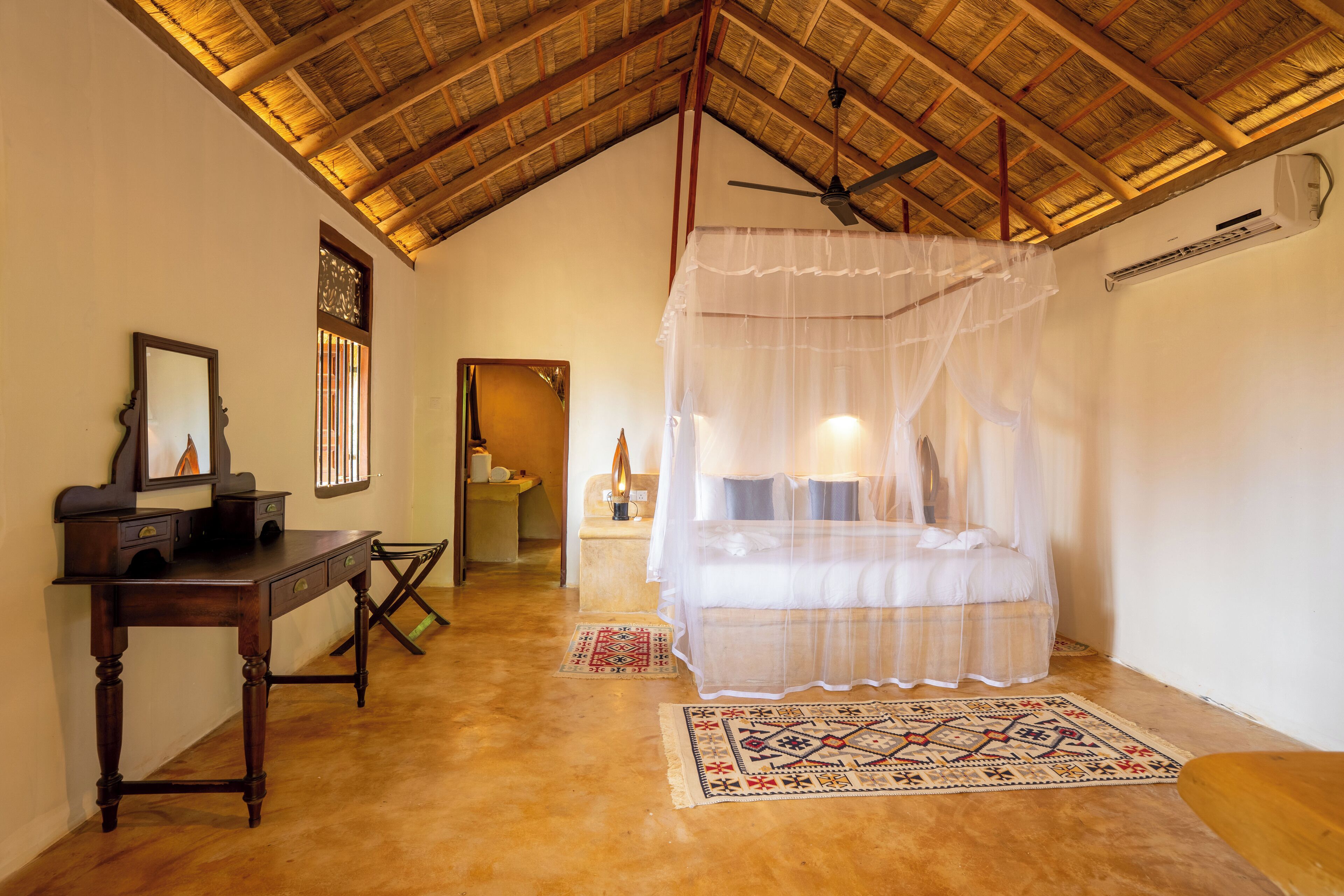 A bedroom with a wooden ceiling, a bed with a white canopy, a desk with drawers, and a small rug.