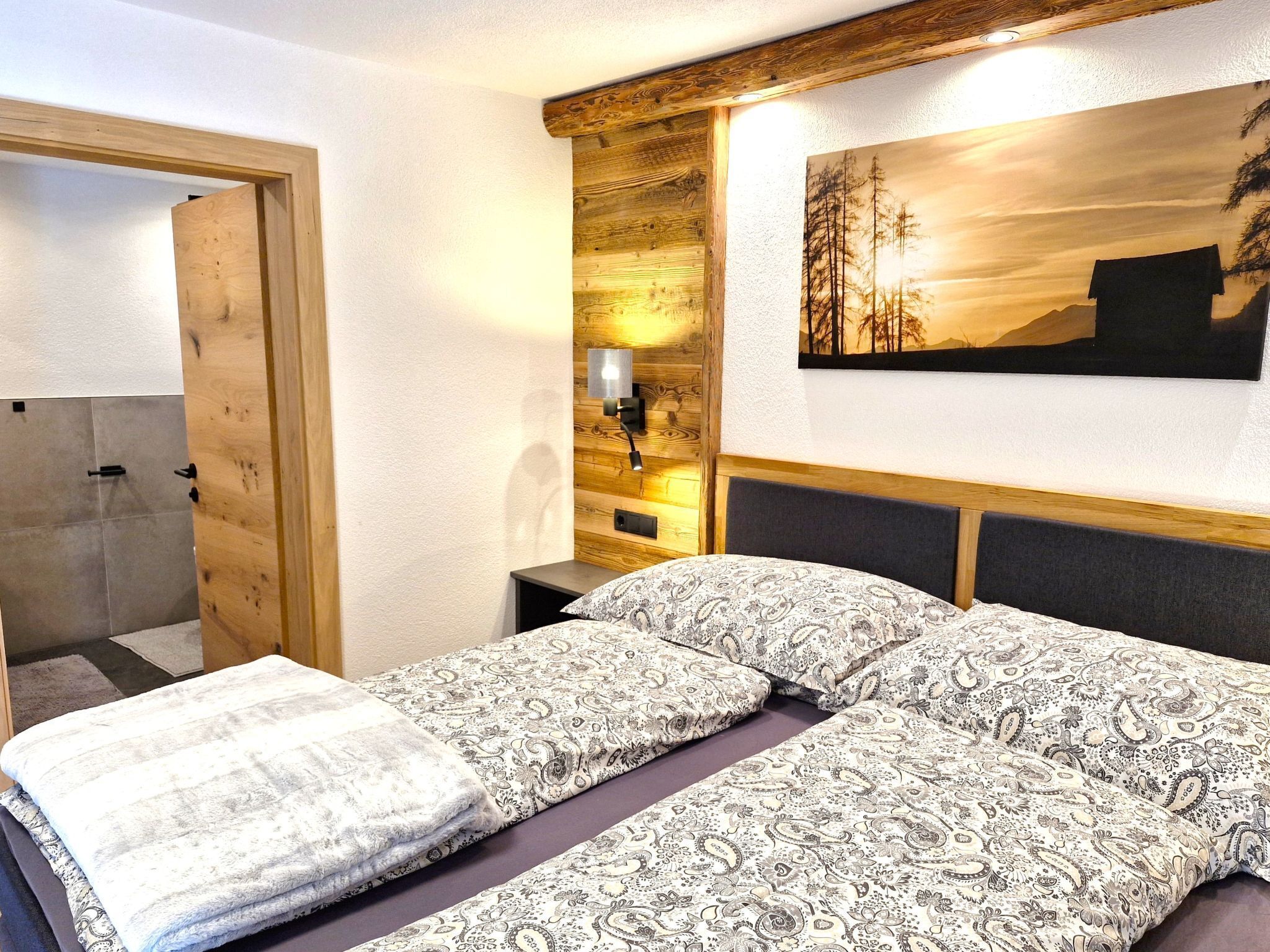 A bedroom with a wooden headboard, a bed with patterned bedding, a nightstand with a lamp, and a painting of a forest scene.
