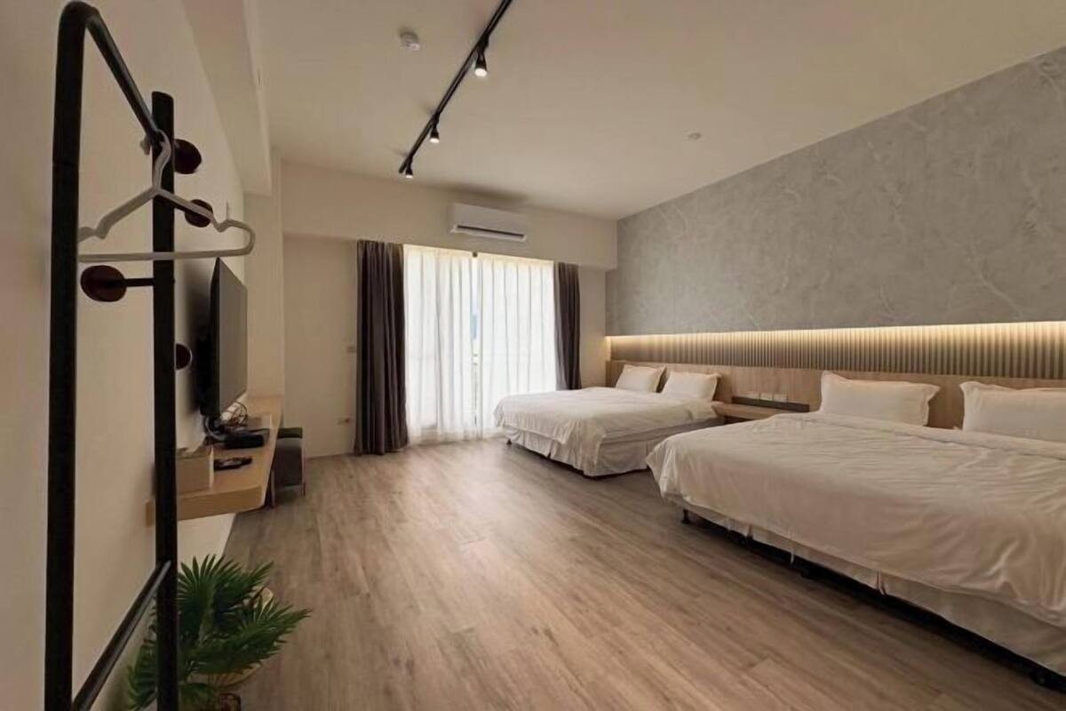 A modern hotel room with two beds, a wooden floor, a large window with curtains, and a mounted TV.