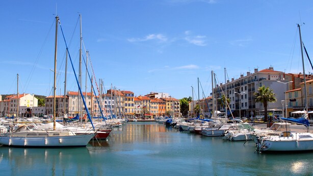 Un port de plaisance où sont amarrés de nombreux bateaux, entouré d’immeubles dotés de balcons et de toits recouverts de tuiles rouges.