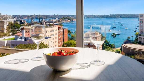 Una mesa con un cuenco de fruta y dos copas de vino, con vistas a un puerto deportivo con barcos y edificios.