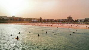 Una playa con gente surfeando y nadando, edificios al fondo y un cielo despejado.