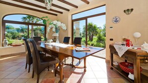 A dining area with a wooden table, chairs, and a vase of white flowers. There are two windows providing a view of the outdoors. - Villa Athena with private pool - My Rental Homes (Castelvetrano)