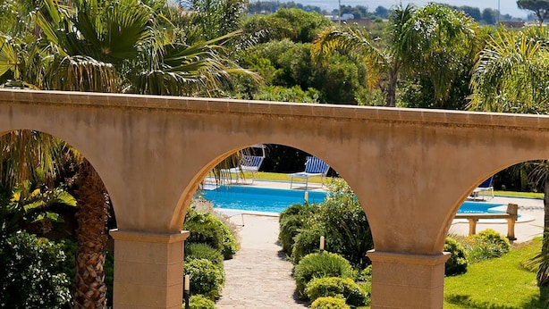A stone bridge with arches leading to a pool area surrounded by lush greenery and palm trees. - Villa Athena with private pool - My Rental Homes (Castelvetrano)