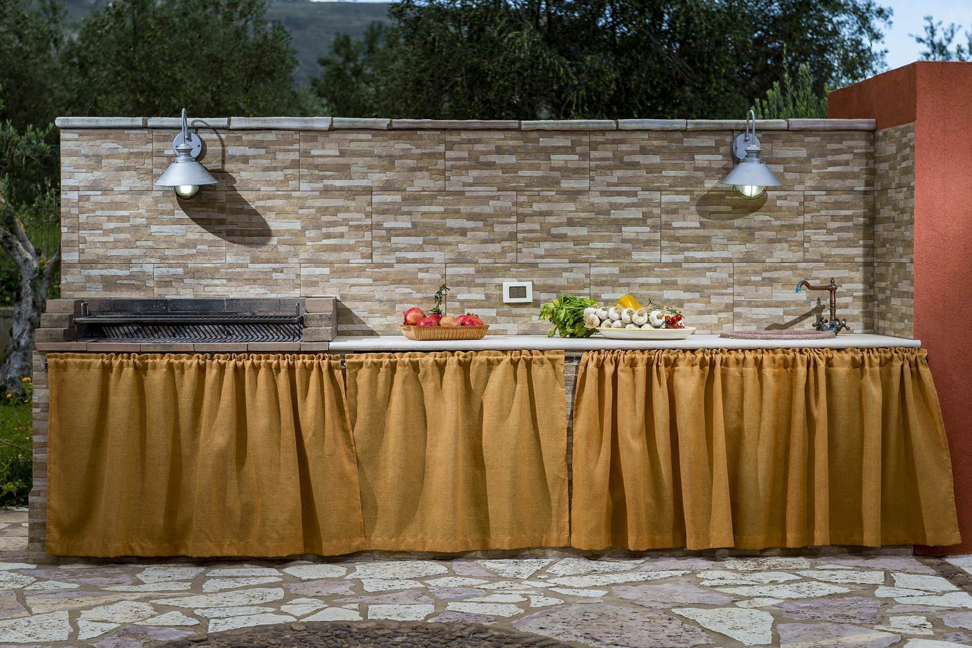 An outdoor kitchen with a stone wall, two pendant lights, a grill, and a table with fresh produce.