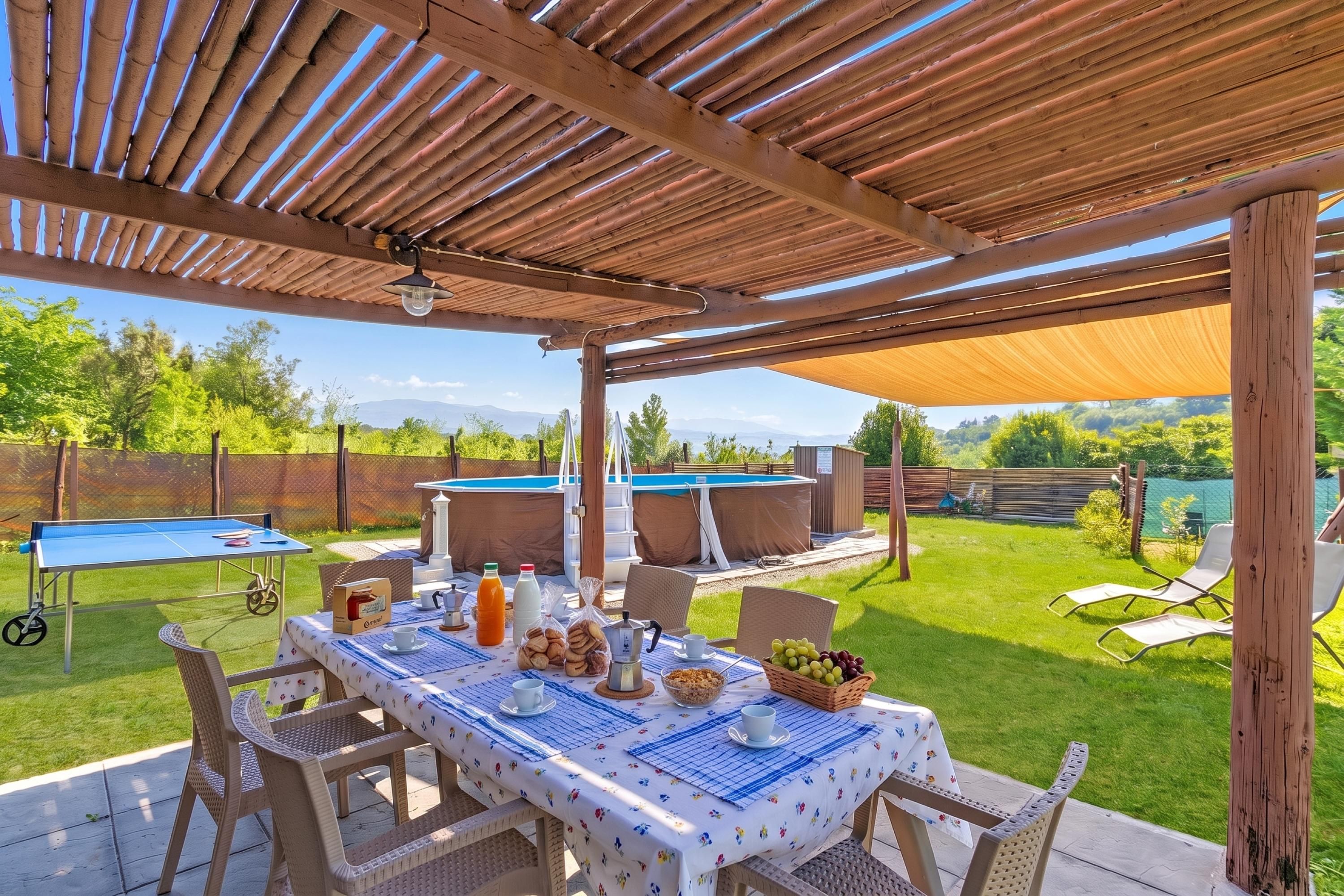 Een terras buiten met een eettafel, omringd door stoelen, een zwembad en een overdekt gedeelte met uitzicht op het landschap.