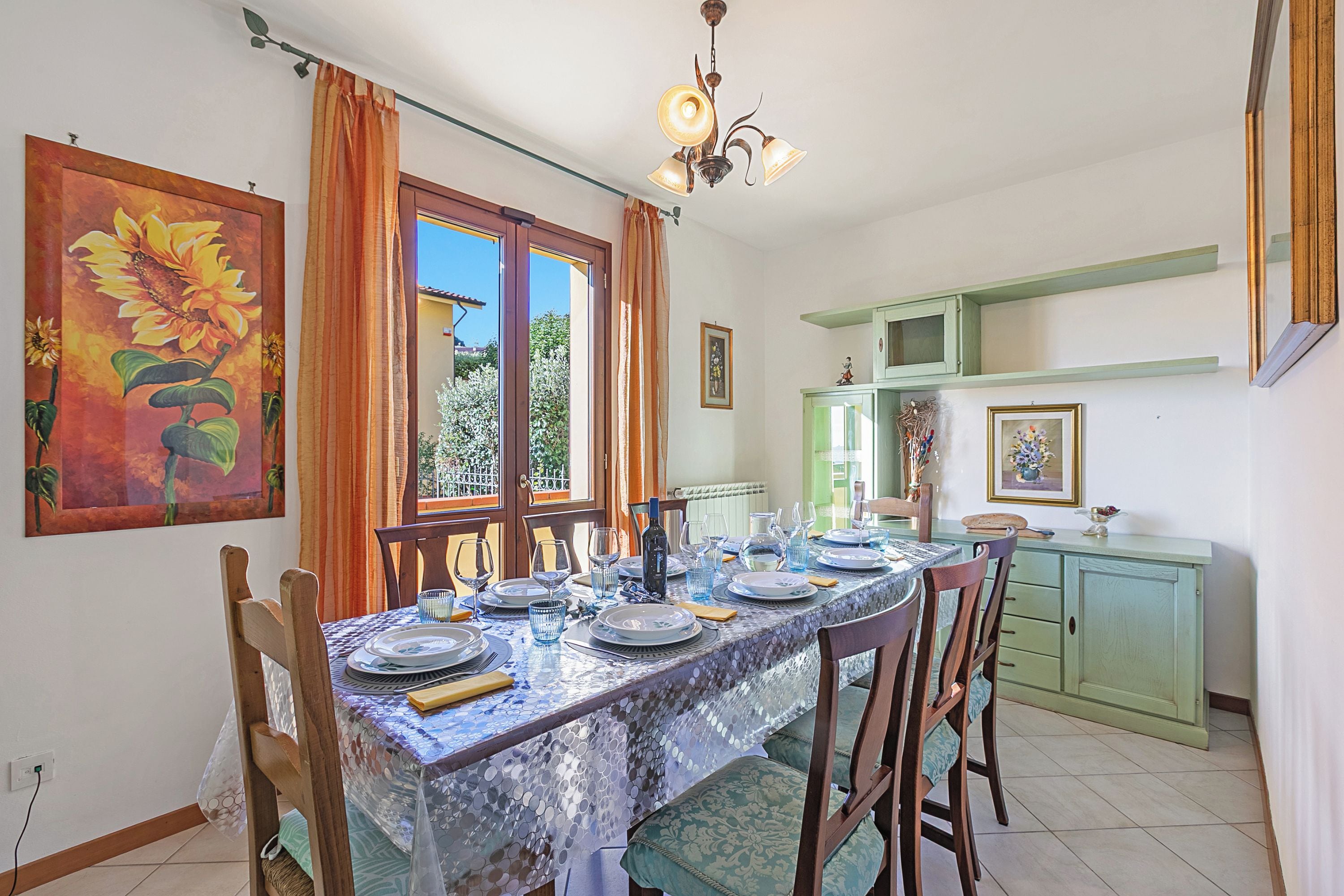 A dining area with a table set for four, a painting of sunflowers on the wall, and a view of greenery outside.