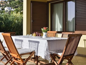 Una mesa al aire libre con mantel blanco, sillas de madera y un jarro de flores.