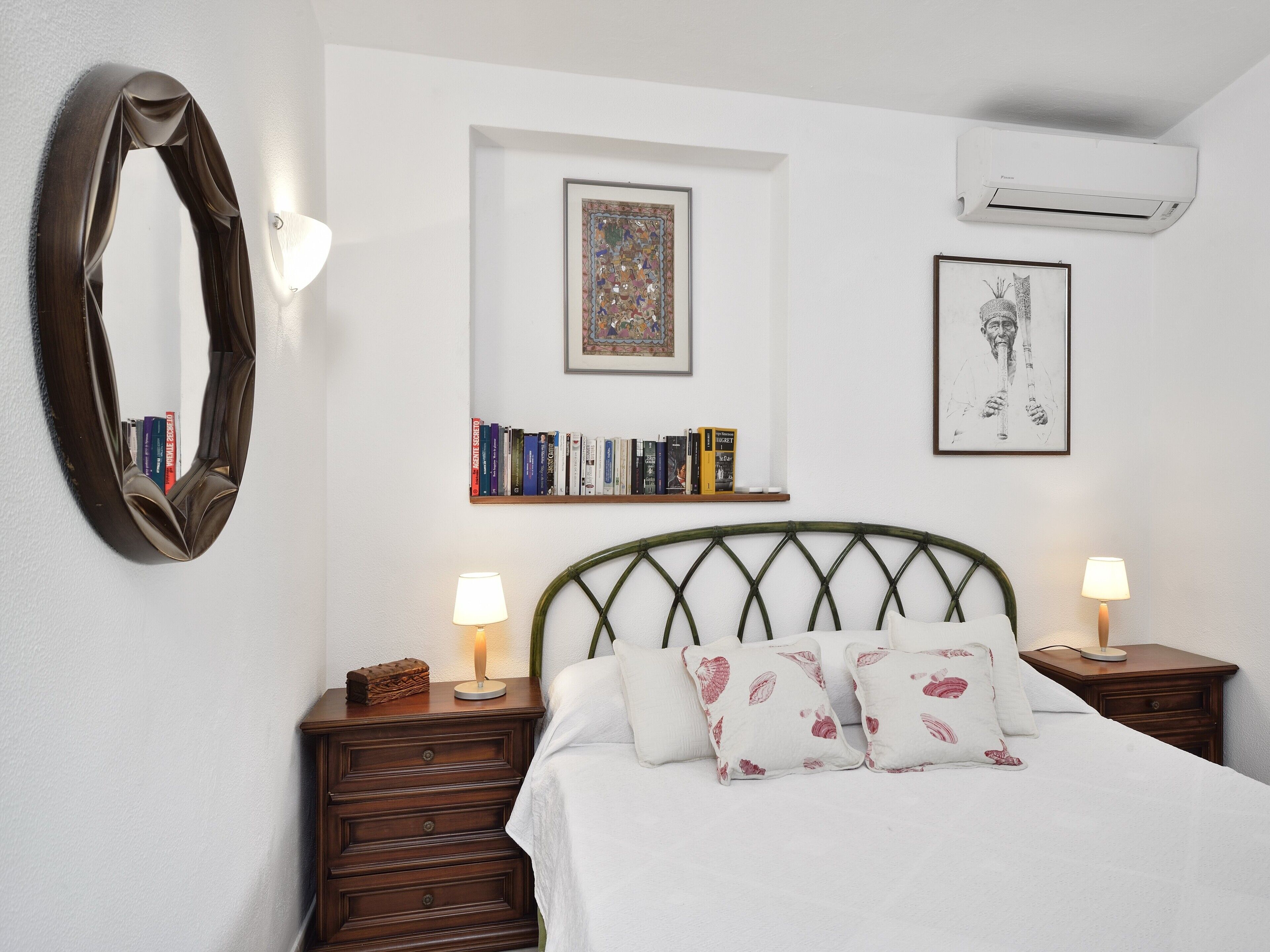 A bedroom with a bed, two bedside tables, a wall-mounted mirror, a bookshelf, and an air conditioning unit.