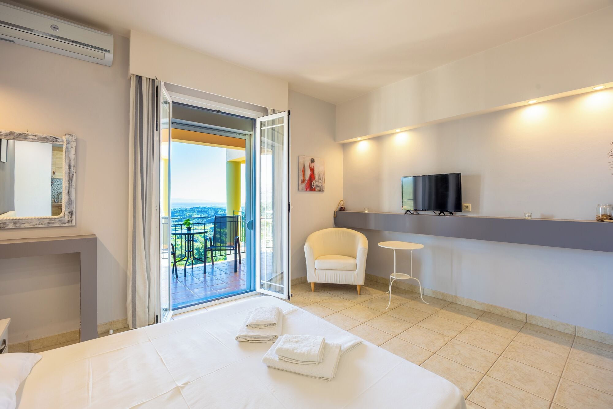 Habitación de hotel con cama, televisor, una silla, una mesita y balcón con vistas.